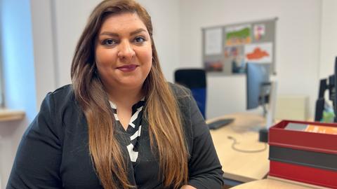 Eine Frau mit schwarzem Blazer und langen, braunen Haaren sitzt an einem Schreibtisch.