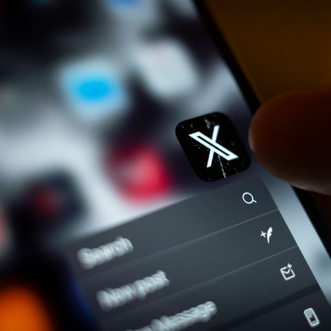 Jemand tippt mit dem Finger die App "X" auf dem Smartphone an. 