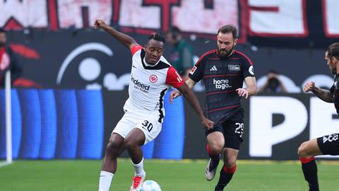 Michy Batshuayi (links im Bild im weißen Eintracht-Trikot mit roten Ärmeln) dribbelt den Ball mit dem linken Fuß. Er wird verfolgt von Lucas Tousart (rechts im Bild, im schwarzen Union-Trikot mit dünnen, roten Querstreifen). 