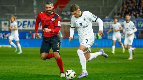 Links im Bild läuft Sascha Mockenhaupt (SV Wehen Wiesbaden, rotes Trikot, schwarze Hose). Er verfolgt Julian Guttau (TSV 1860 München, ganz in weiß) der mit dem Ball dribbelt (rechts im Bild). 