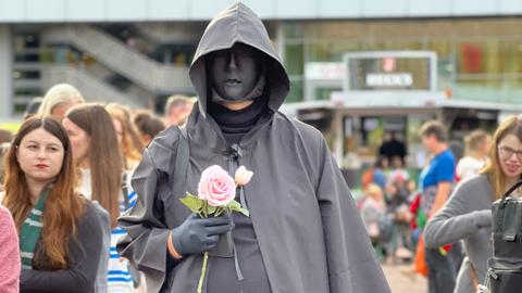 Das Bild zeigt einen Cosplayer auf der Buchmesse 2024. Er trägt einen schwarzen Umhang mit Kapuze und eine schwarze Gesichtsmaske. In der Hand hält er eine rosafarbene Rose.