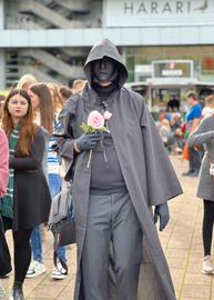 Das Bild zeigt einen Cosplayer auf der Buchmesse 2024. Er trägt einen schwarzen Umhang mit Kapuze und eine schwarze Gesichtsmaske. In der Hand hält er eine rosafarbene Rose.