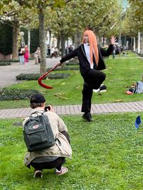 Das Bild zeigt Cosplayer auf der Buchmesse 2024. Eine Frau mit einer pfirsichfarbenen Langhaarperücke und schwarzem Anzug posiert für einen Mann mit Rucksack, der auf einer Wiese kniet und sie fotografiert.