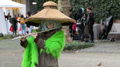 Das Foto zeigt eine Cosplayerin auf der Buchmesse 2024. Sie trägt einen übermäßig großen Strohhut und hält eine Querflöte vor den Mund.