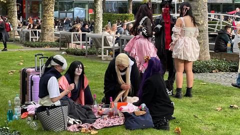 Das Bild zeigt eine Gruppe Cosplayer auf der Buchmesse 2024, die auf einer Picknickdecke auf einer Grünfläche auf dem Messegelände sitzen.