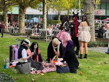 Das Bild zeigt eine Gruppe Cosplayer auf der Buchmesse 2024, die auf einer Picknickdecke auf einer Grünfläche auf dem Messegelände sitzen.