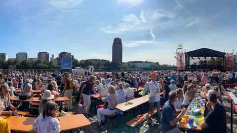 Das Bild zeigt das Gelände an der Weseler Werft in Frankfurt. Menschen mit Sonnenbrillen und Sonnenhüten sitzen auf Bierbänken, im Hintergrund sind der Main und eine leere Bühne zu sehen.