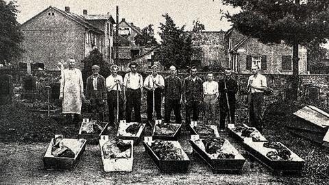 Historisches s/w-Bild mit Menschen, die auf einer Straße hinter geöffneten Särge stehen.