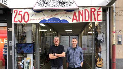 Zwei Männer stehen vor einem Ladengeschäft und schauen in die Kamera. Über ihnen ein Scild, auf dem steht: "250 Musikhaus André"