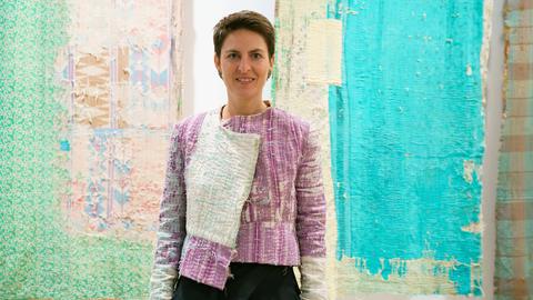 Anna Heringer in einem rosa-weißen Patchwork-Jackett vor Patchwork-Arbeiten in Türkis, Rosa und Beige