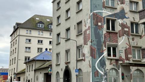 Das Bild zeigt das Atelier Frankfurt von außen. Zu sehen sind zwei graue Gebäude. Auf die Fassade des vorderen Gebäudes sind Mauerstücke aus rotem Stein sowie Buchstaben und blaue Drachen-artige Tiere gemalt worden.