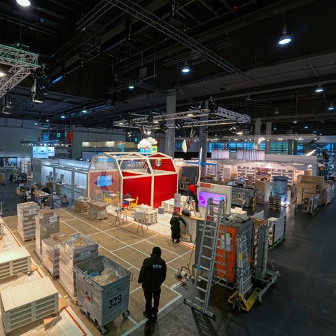 Das Bild zeigt ein Messehalle im Aufbau. Im Raum stehen silberne Leitern, Holzkisten und Paletten. Auch halbfertige Messestände sind bereits zu erkennen.