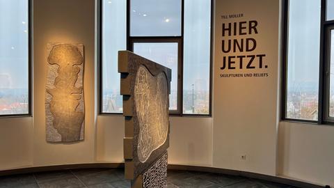 Eine große, runde Ausstellungshalle. Zum Teil sind alte Backsteine zu sehen. Im Mittelpunkt sind Kunstwerke aus Holz und Stein. Dort sind Worte und Zeichnungen eingeritzt und coloriert.