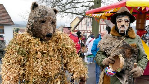 Zwei kostümierte Menschen schauen in die Kamera: links ein Bär aus Stroh, rechts ein Mann mit Filzhut und Tamburin in der Hand.