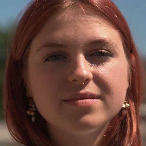 Das Bild zeigt eine junge Frau mit rot gefärbten Haaren und T-Shirt. Sie hat silberne Over-Ear-Kopfhörer um den Hals. Im Hintergrund ist ein Park zu sehen.