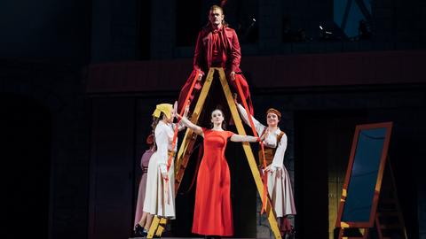 Das Bild zeigt eine Szene aus dem  Musical "Gänsemagd". Ein in rot gekleideter Mann sitzt auf einer als "A" aufgestellten Leiter. Auf den untersten Sprossen stehen auf beiden Seiten in weiß gekleidete Frauen. Mittig zwischen den Leiterbeinen steht eine Frau in einem roten Kleid.