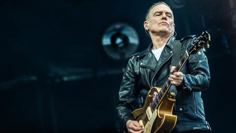 Das Bild zeigt Rockmusiker Bryan Adams. Zu sehen ist ein Mann mit grauen, nach hinten gegelten Haaren. Er trägt eine Lederjacke über einem weißen T-Shirt und spielt Gitarre. Seine Augen sind geschlossen.