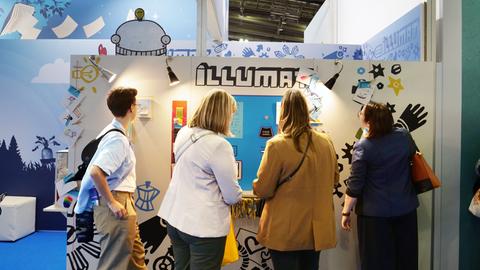 Das Bild zeigt einen Stand auf der Frankfurter Buchmesse. Vier Frauen stehen vor dem sogenannten Illumaten, der optisch wie ein Fotoautomat gestaltet ist. Hinter der Pappwand sitzt ein Illustrator, der in Echtzeit Wunschbilder umsetzt.