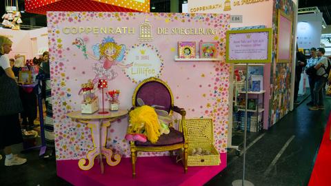 Das Bild zeigt einen Stand auf der Frankfurter Buchmesse. Zu sehen ist ein Prinzessinnenzimmer mit rosa Hintergrund, gold-glitzerndem Spiegel und einem mit rotem Samt bezogenem Thron.