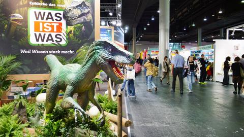 Das Bild zeigt einen Stand auf der Frankfurter Buchmesse. Davor ist ein lebensgroßer grüner Tyrannosaurus Rex aufgestellt.