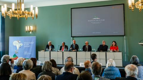 Fünf Männer und eine Frau sitzen auf einem Podium an einem langen Tisch, im Vordergrund Publikum auf Stühlen von hinten