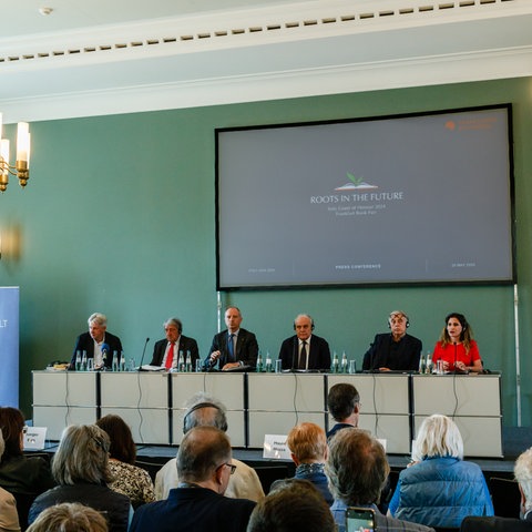 Fünf Männer und eine Frau sitzen auf einem Podium an einem langen Tisch, im Vordergrund Publikum auf Stühlen von hinten