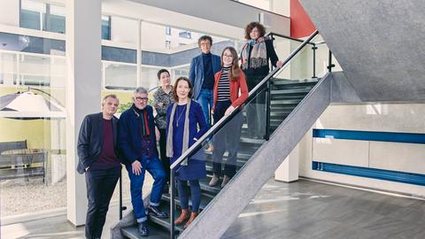 Die sieben Mitglieder der Jury des Buchpreises 2024 stehen hintereinander auf einer Treppe in einer hellen Eingangshalle.