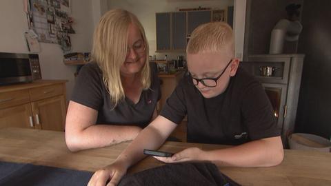 Das Bild zeigt eine blonde Frau mit schulterlangen glatten Haaren und silber gerahmter runder Brille, die gemeinsam mit einem blonden Jungen mit schwarz gerahmter Brille an einem Holztisch sitzt und auf ein Handy schaut, das der Junge in den Händen hält. 