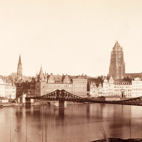 Schwarz-weiß-Foto eines Flusses, über den eine Eisenbrücke führt. Auf der anderen Seite des Flusses steht eine Häuserzeile, aus der zwei Kirchtürme ragen.