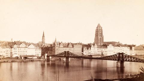 Schwarz-weiß-Foto eines Flusses, über den eine Eisenbrücke führt. Auf der anderen Seite des Flusses steht eine Häuserzeile, aus der zwei Kirchtürme ragen.