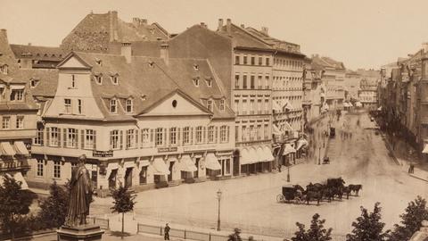 Schwarz-weiß-Foto einer Straßengabelung, an der Häuser und ein Platz liegen. Auf der Straße fährt eine Kutsche. Auf dem Paltz steht eine umzäunte Statue, die quasi auf die Weggabelung blickt.