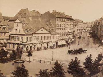 Schwarz-weiß-Foto einer Straßengabelung, an der Häuser und ein Platz liegen. Auf der Straße fährt eine Kutsche. Auf dem Paltz steht eine umzäunte Statue, die quasi auf die Weggabelung blickt.