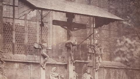 Schwarz-weiß-Foto eines überdachten Kreuzes vor einer Kirche. Davor stehen mehrere kleine Statuen.