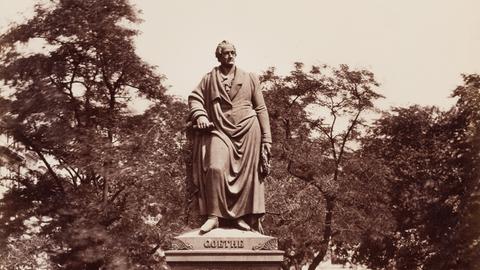 Schwarz-weiß-Foto der Statue eines Mannes auf einem quadratischen Sockel. Die Statur ist umzäunt und steht auf einem Platz vor einer Reihe von Bäumen.