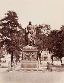 Schwarz-weiß-Foto der Statue eines Mannes auf einem quadratischen Sockel. Die Statur ist umzäunt und steht auf einem Platz vor einer Reihe von Bäumen.