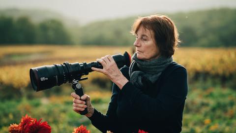 Eine Frau Steht in einem Feld. In der rechten Hand hält sie eine Kamera mit langem Objektiv und schaut darauf. Sie hat halblange braune Haare und trägt einen dunklen Pullover und einen grauen Schal. Der Hintergrund ist unscharf.