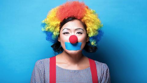 Das Bild zeigt eine als Clown verkleidete und geschminkte Frau. Sie trägt eine lockige Perücke in rot, gelb und blau über ihren dunklen halblangen Haaren. Über ihrem Mund klebt ein blaues Stück Klebeband.