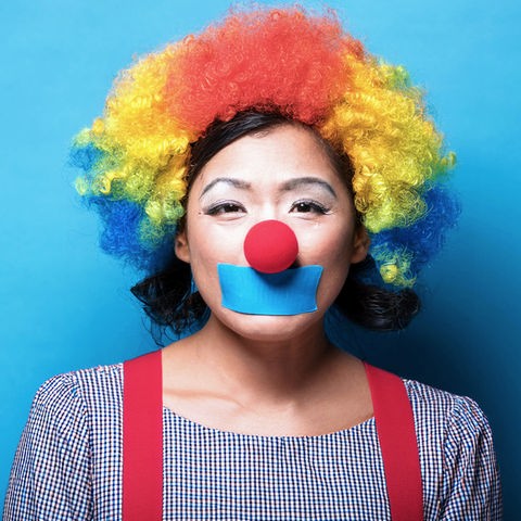 Das Bild zeigt eine als Clown verkleidete und geschminkte Frau. Sie trägt eine lockige Perücke in rot, gelb und blau über ihren dunklen halblangen Haaren. Über ihrem Mund klebt ein blaues Stück Klebeband.