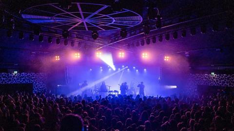 Band auf einer Bühne, die in violett-blaues Licht getaucht ist. Im Dunkeln um die Bühne viele Konzertbesucher