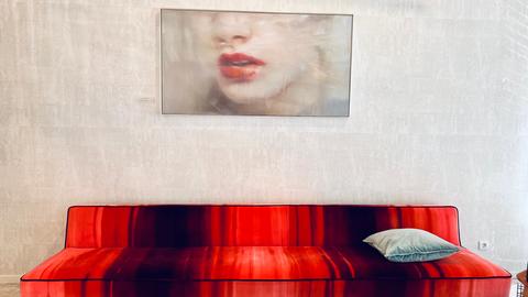 Das Bild zeigt ein Gemälde, darunter ein rotes Sofa mit Sitzkissen. Das Gemälde zeigt das Gesicht einer Frau von der Nase abwärts. Sie hat rot geschminkte Lippen und den Mund leicht geöffnet.