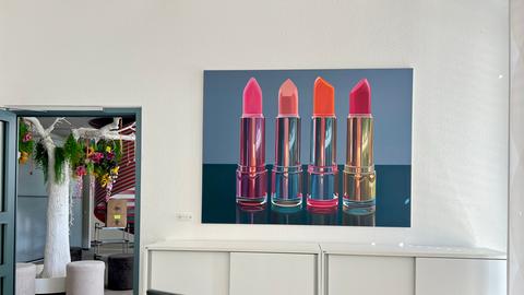 Das Bild zeigt eine großformatige Fotografie von drei verschiedenfarbigen Lippenstiften in einem Büro.