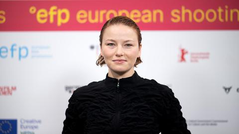 Eine junge Frau (Oberkörperportrait) blickt in die Kamera. Im Hintergrund unscharf eine Wand mit vielen Logos und dem Schriftzug "efp european shooting..."