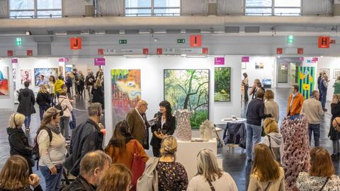 Das Bild zeigt das Messegeschehen auf der Kunstmesse Discovery Art Fair in Frankfurt. Zu sehen sind verschiedene Menschen, die vor einem Gemälde stehen.