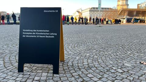 Vor einem großen Gebäude mit einer Glasfassade warten unzählige Menschen auf den Einlass