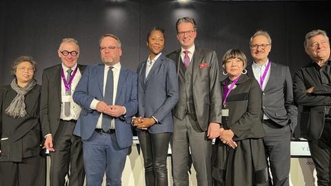 Pressekonferenz zur neuen künstlerischen Leitung der documenta 16: Eine Gruppe von drei Frauen und fünf Männern steht nebeneinander. Sie tragen formelle Kleidung und lächeln in die Kamera. Einige tragen lila Bänder mit Schildchen um den Hals.