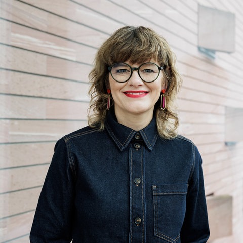 Eine Frau mit schulterlangem, lockigem Haar steht vor einer modernen Gebäudefassade. Sie trägt eine runde, schwarze Brille, leuchtend roten Lippenstift, auffällige Ohrringe und ein dunkelblaues Jeanshemd. Im Hintergrund sind unscharf eine mittelalterliche Stadtansicht mit Türmen und Dächern zu erkennen.