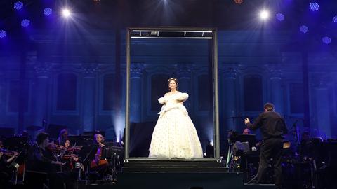 Eine Frau in einem eleganten, weißen, bauschigen Ballkleid steht in einem beleuchteten Rahmen auf der Bühne. Im Hintergrund ist ein Orchester zu sehen, dessen Mitglieder verschiedene Instrumente spielen. Die Bühne ist in blauem Licht getaucht, und Scheinwerfer strahlen von oben.