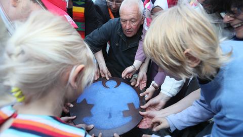 Ein Mann und mehrere Kinder halten eine Scheibe, die einen Kreis symbolisiert, in den ein Halbmond, ein Kreuz und ein Davidstern eingearbeitet sind. Die Auslassungen zeigen einen Engel.