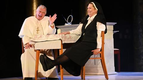 Papst und Nonne unterhalten sich lachend an einem Tisch.