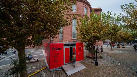 Das Bild zeigt einen rot verkleideten Container auf dem Paulsplatz in Frankfurt. Es handelt sich um einen Escape Room zur Aktion "The Great Escape Room – Eine inspirierende Auszeit auf den Spuren Goethes Italienischer Reise". Im Hintergrund ist die Paulskirche aus rotem Sandstein zu sehen. 